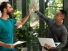 Zwei junge Männer stehen in einem modernen Büro mit viel Grün und geben sich ein motiviertes High-Five; einer hält ein Laptop, der andere ein Blatt Papier. Das Bild symbolisiert erfolgreiche Zusammenarbeit und Teamgeist.